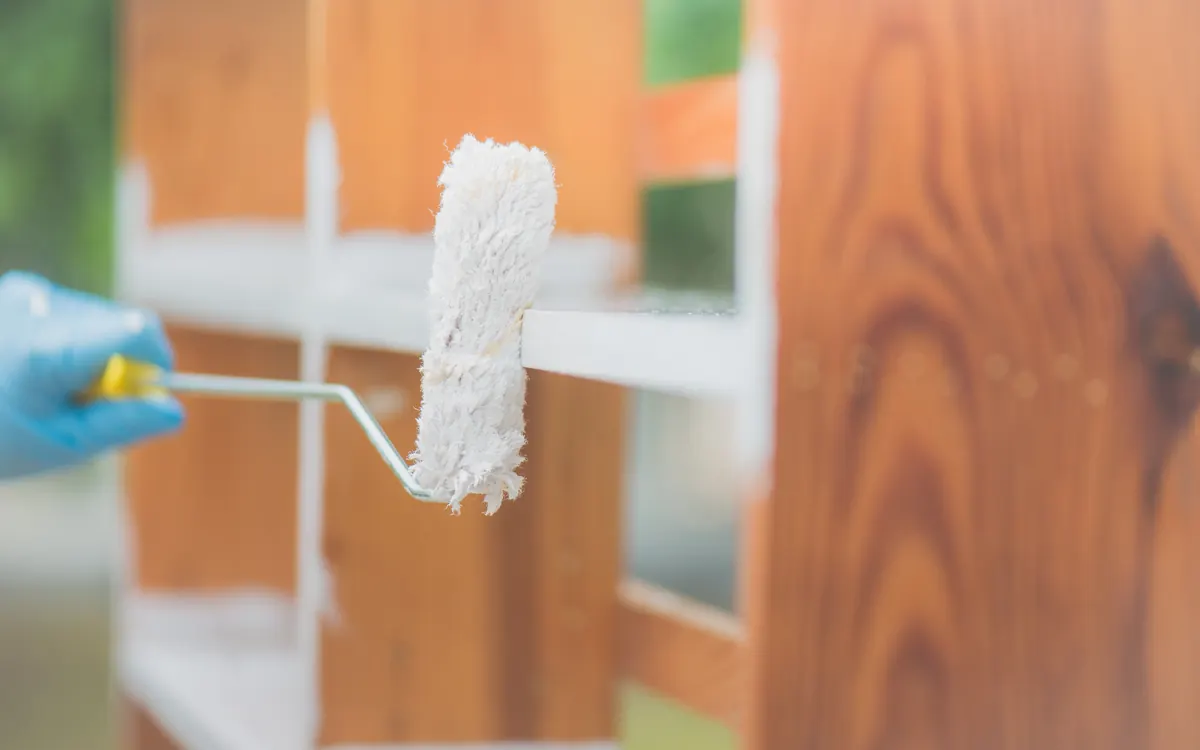 Roller applying white paint to a stained wood cabinet frame