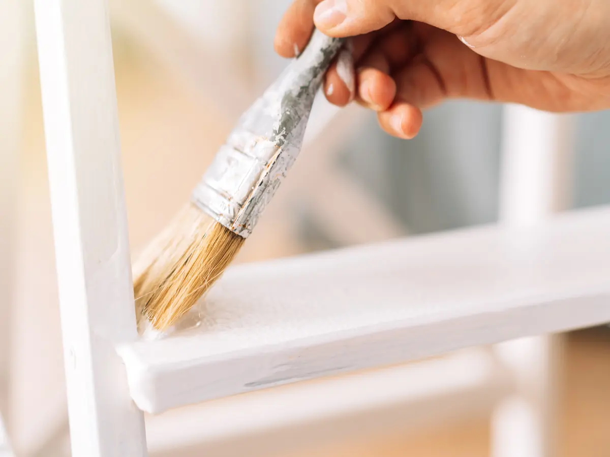 Brush applying white paint to a wooden furniture frame