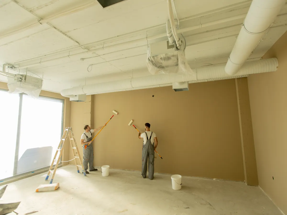 Two painters rolling fresh paint onto the walls of an empty room with drop cloths down