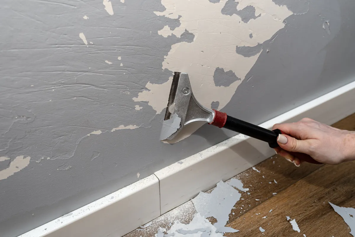 Scraper removing old wallpaper and paint layers from a wall above white baseboard, wallpaper removal by Blue Oak Painting Company in Houston, TX